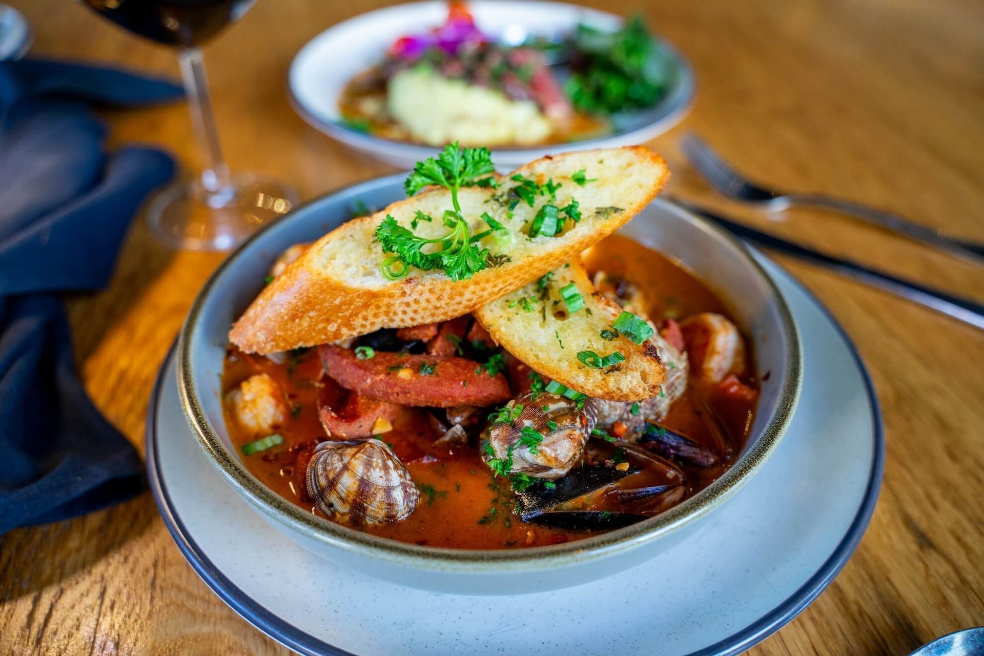 Seafood stew with toasted bread garnish in a bowl, plus another dish and red wine in background.