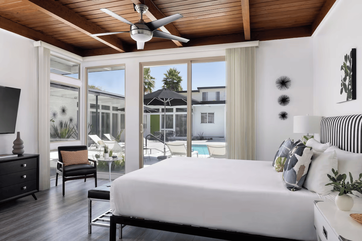 Bright bedroom with a white bed, open doors to a pool patio, and a wood-paneled ceiling with a fan