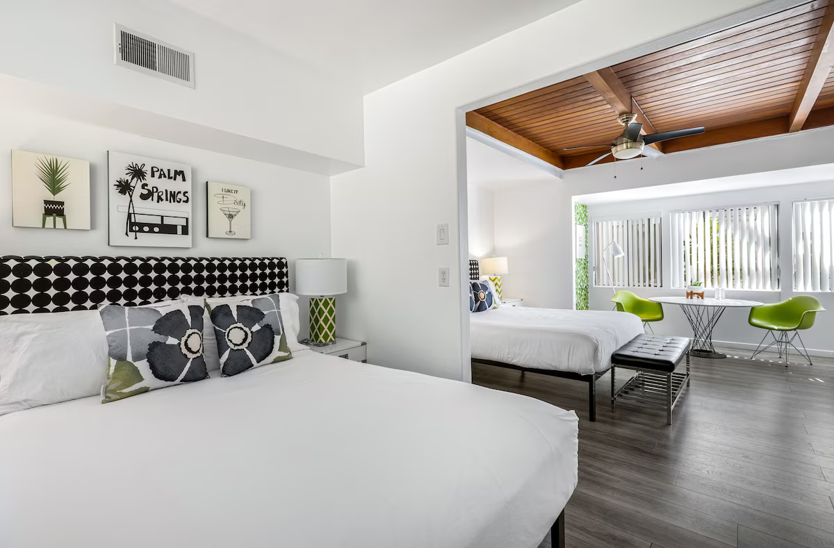 Bright modern bedroom with white bed, patterned pillows, and an adjoining lounge with green chairs.
