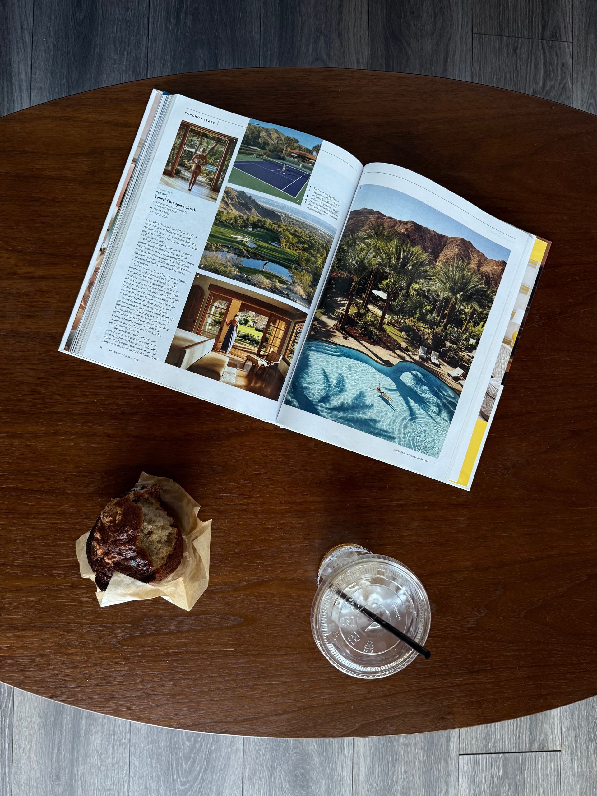 Open magazine on a wooden table beside a dessert and a drink with a straw.