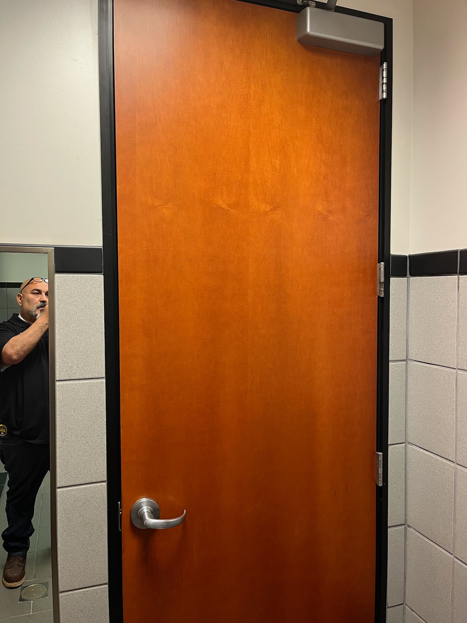 Brown door with a lever handle and automatic door closer; person reflected in a mirror.