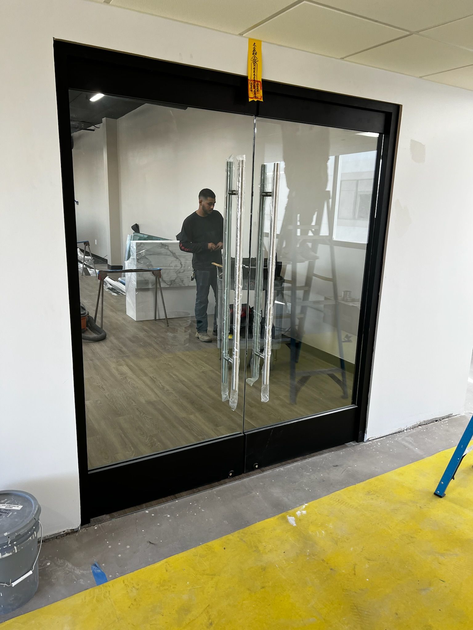 Glass door with black frame reflecting an office interior and a person. Yellow floor, white walls.