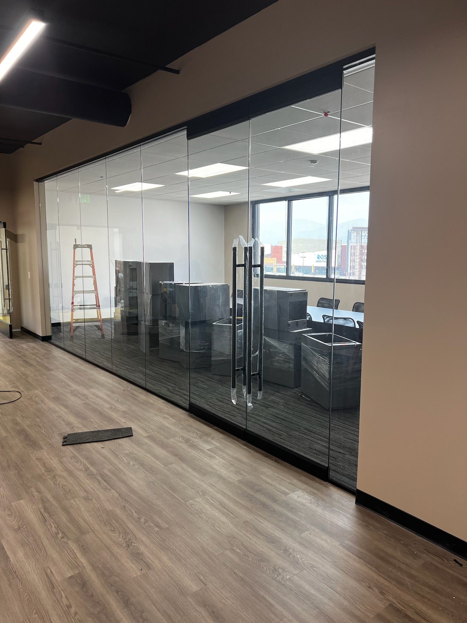 Glass-walled office space with conference table and chairs visible. Light wood floor, neutral walls, black ceiling.