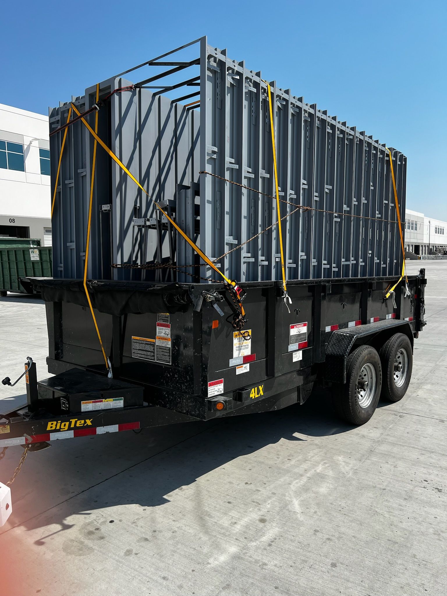 Black dump trailer with a gray cage, secured by yellow straps, parked outside on a sunny day.