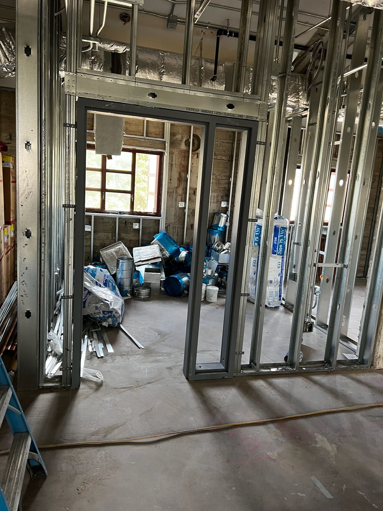 Interior view of a room under construction with metal framing and an open doorway; materials are piled in the doorway.