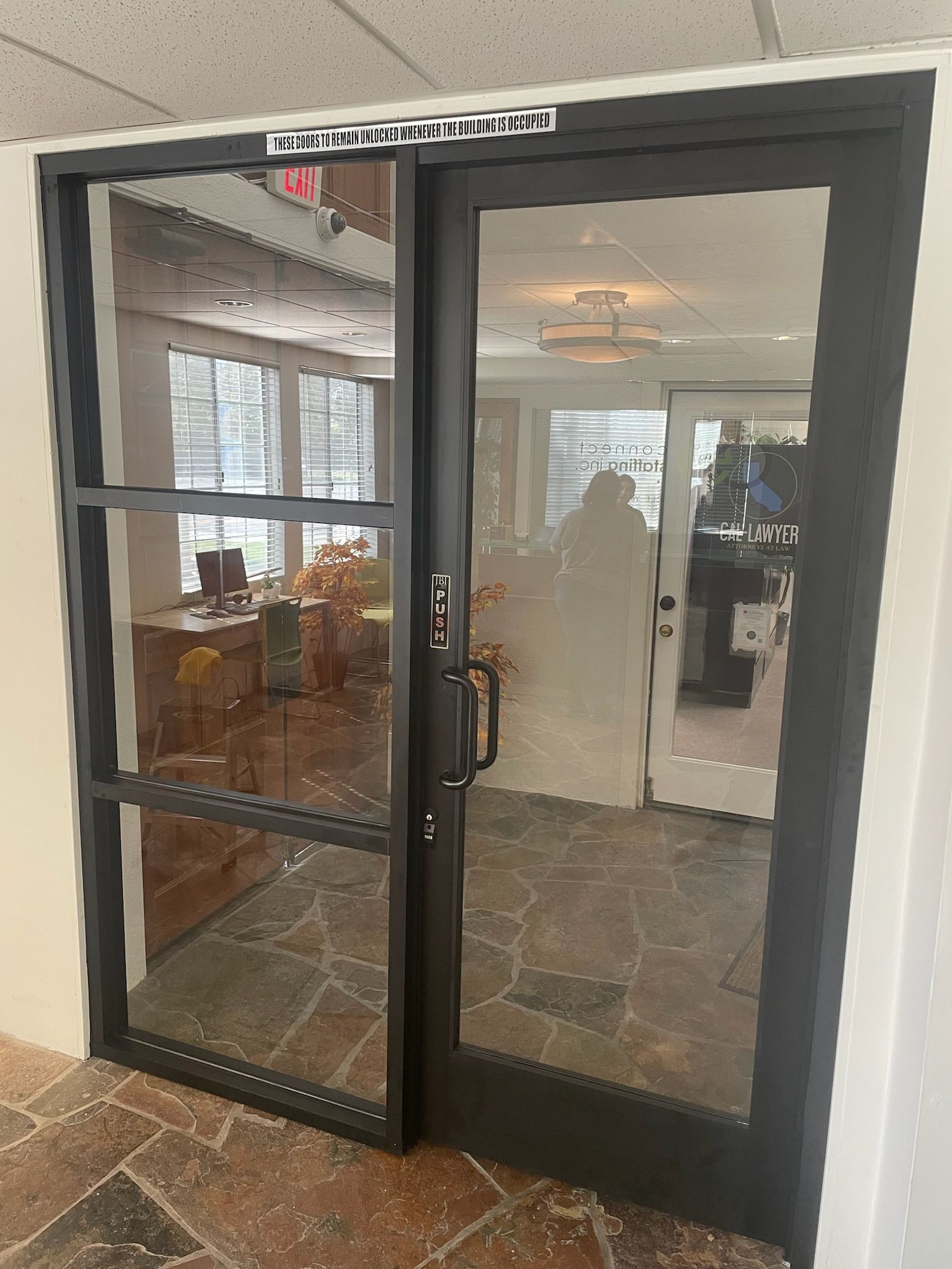 Black-framed glass doors, one sliding, leading into a room with office furniture and natural light.