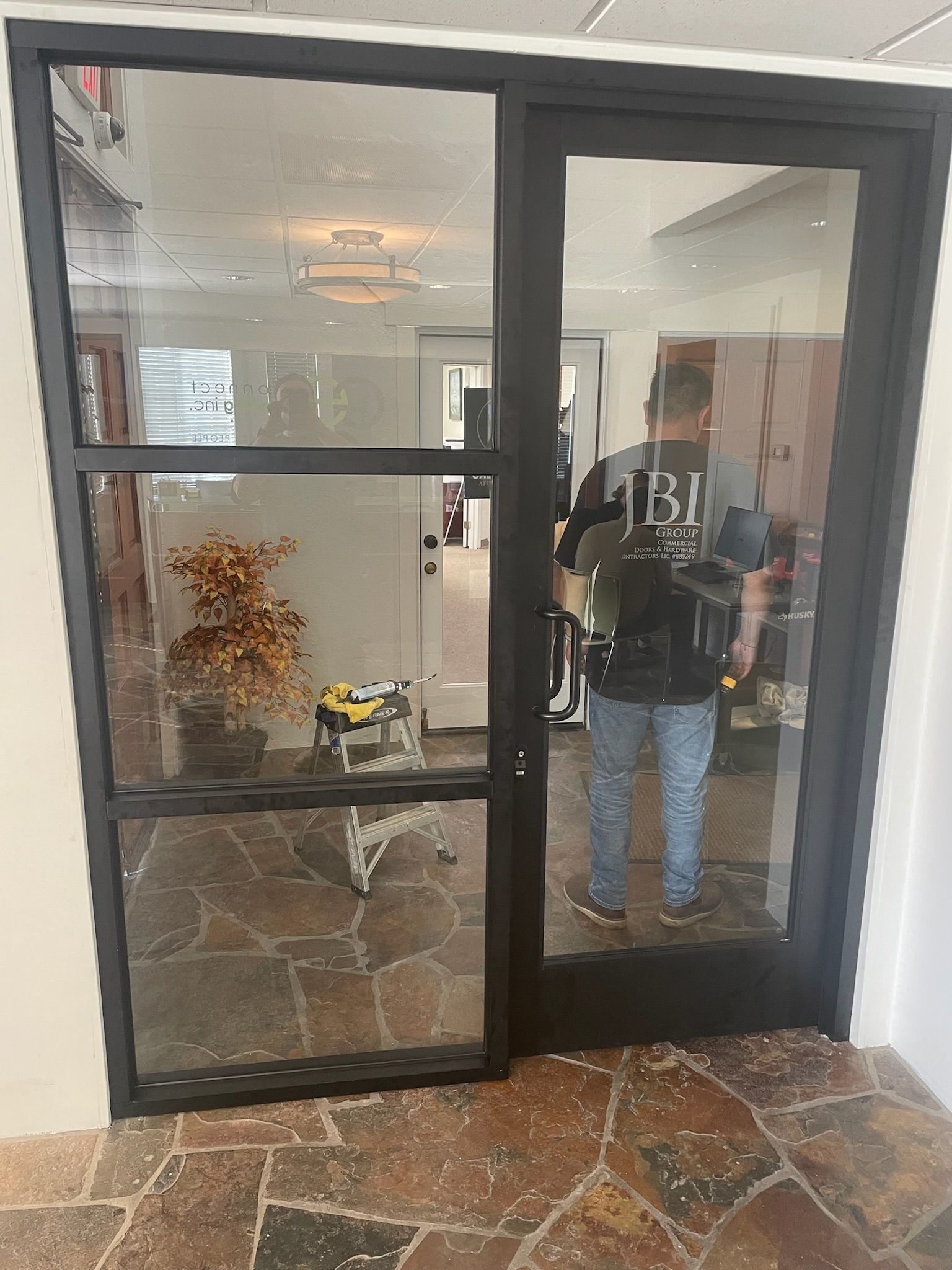 Black framed glass door, person holding handle, interior hallway visible.