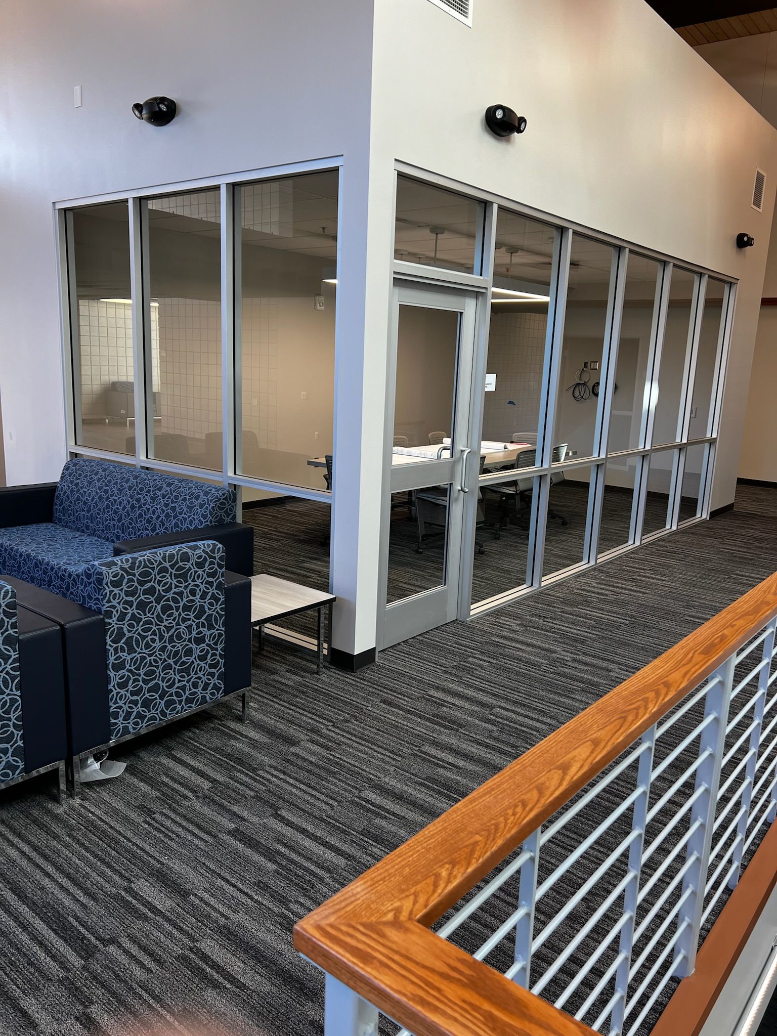 Lounge area with blue patterned seating, glass-walled office, and a wooden railing.
