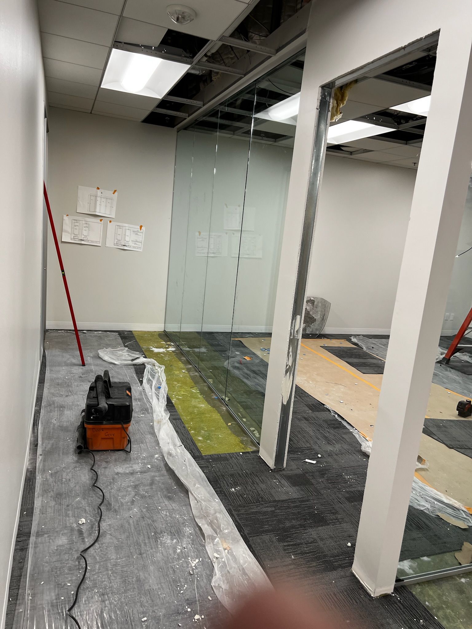 Office construction site. Walls, partially installed flooring, exposed ceiling, power tool, and glass panels visible.