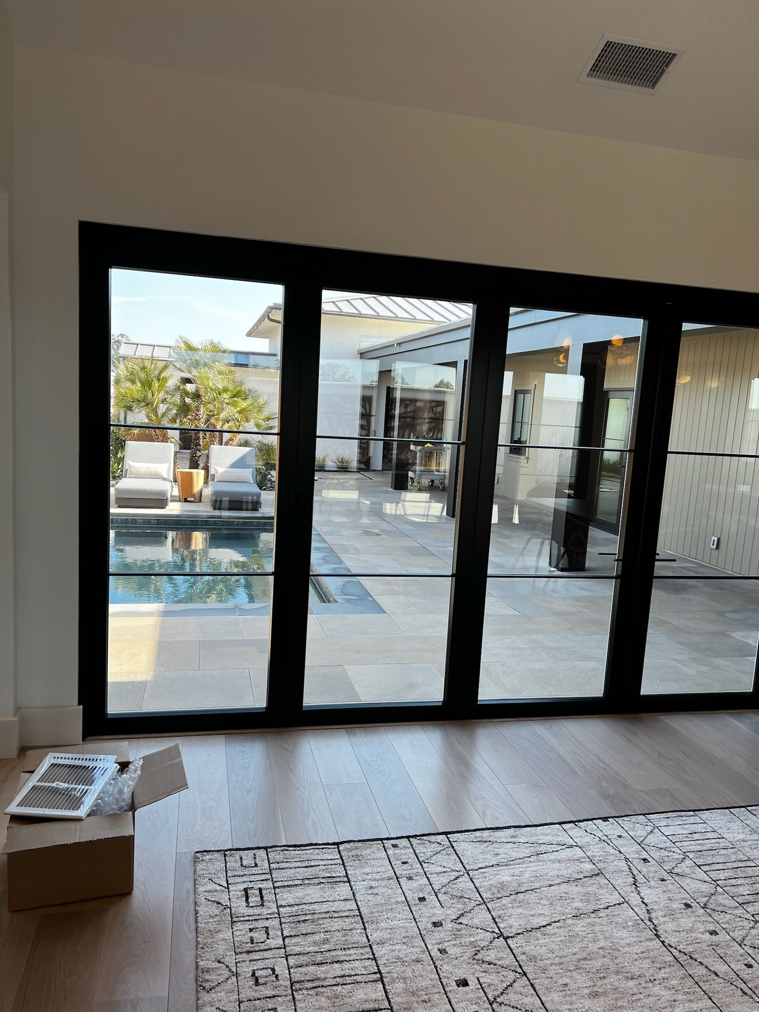 Black framed glass doors open to a patio with pool. Hardwood floor, rug, and cardboard box in the room.