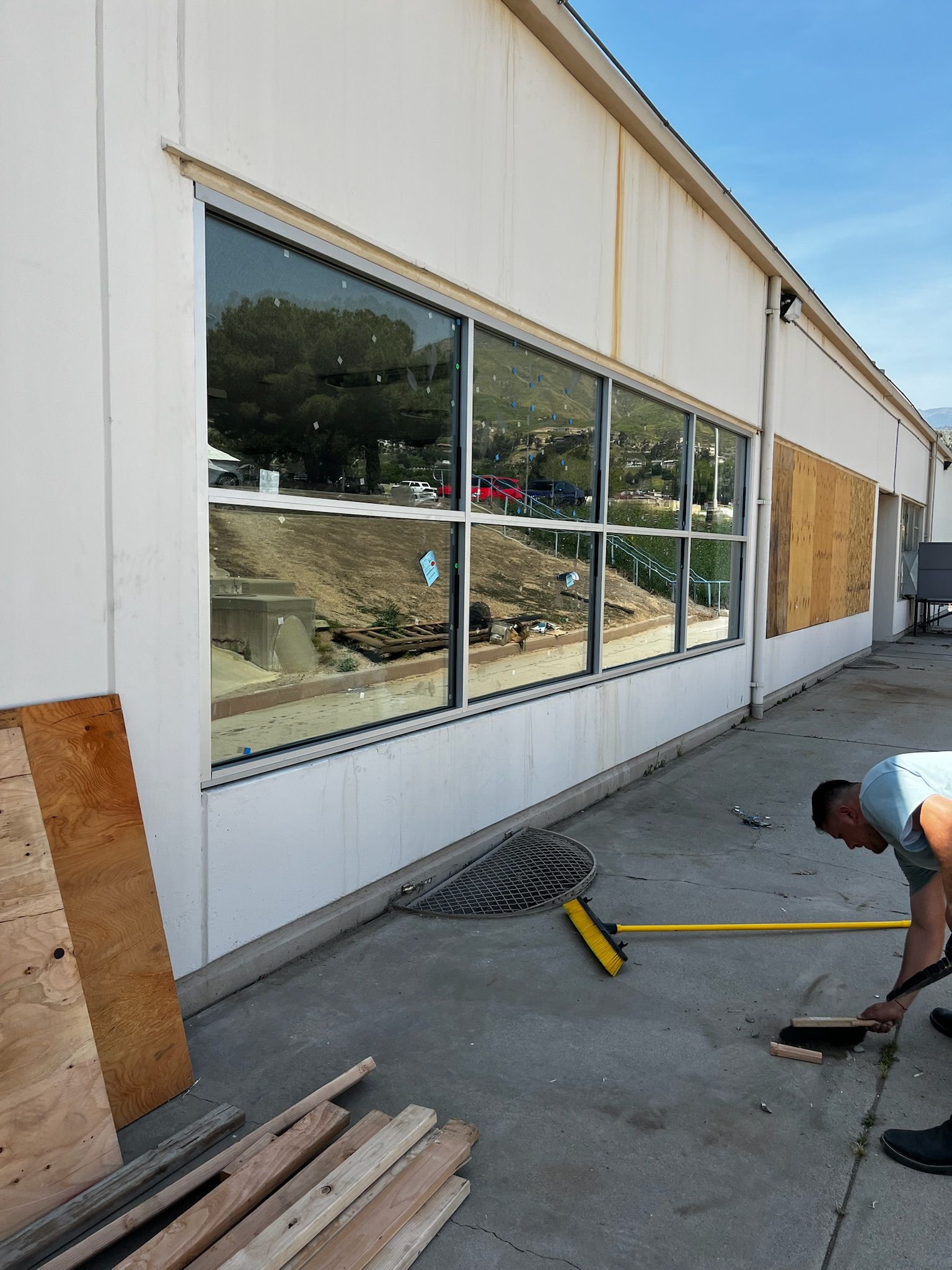 Exterior of a building with a man sweeping; boarded-up windows and large window reflecting a construction site.