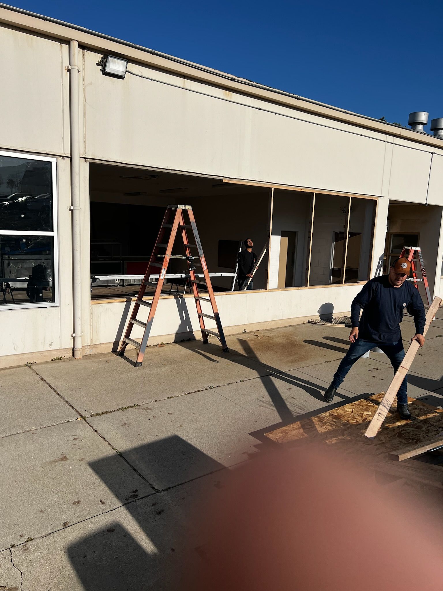 Person wielding a wooden plank outside a building with an open window; ladder visible.