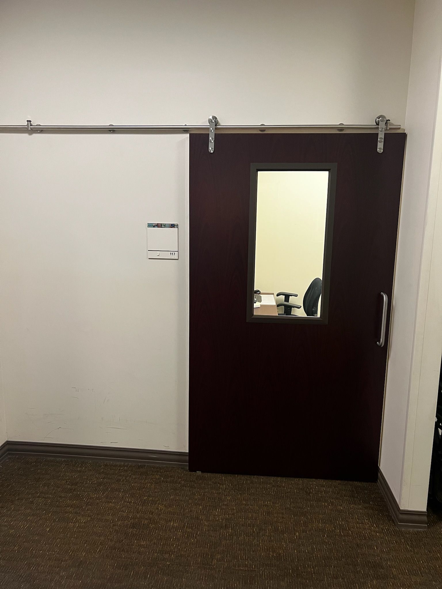Barn door with a rectangular window, burgundy, on a gray rail. It leads to an office.