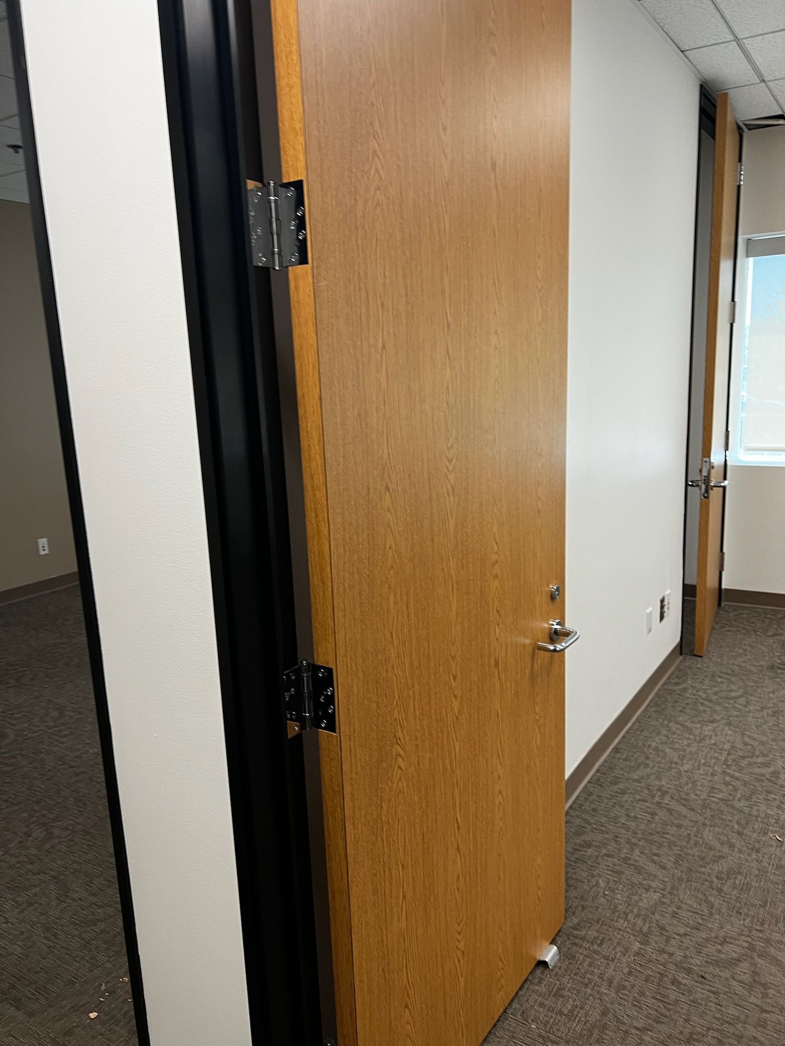 Oak-colored office door slightly ajar, revealing a hallway and the interior of a room with carpet.
