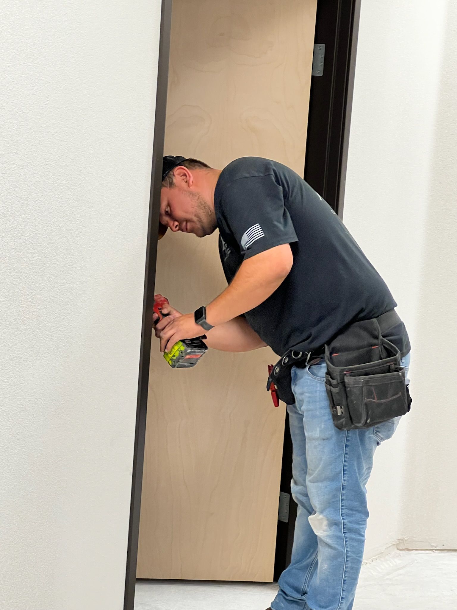 Man using a drill on a door, likely installing it. He wears a tool belt and jeans, indoors.