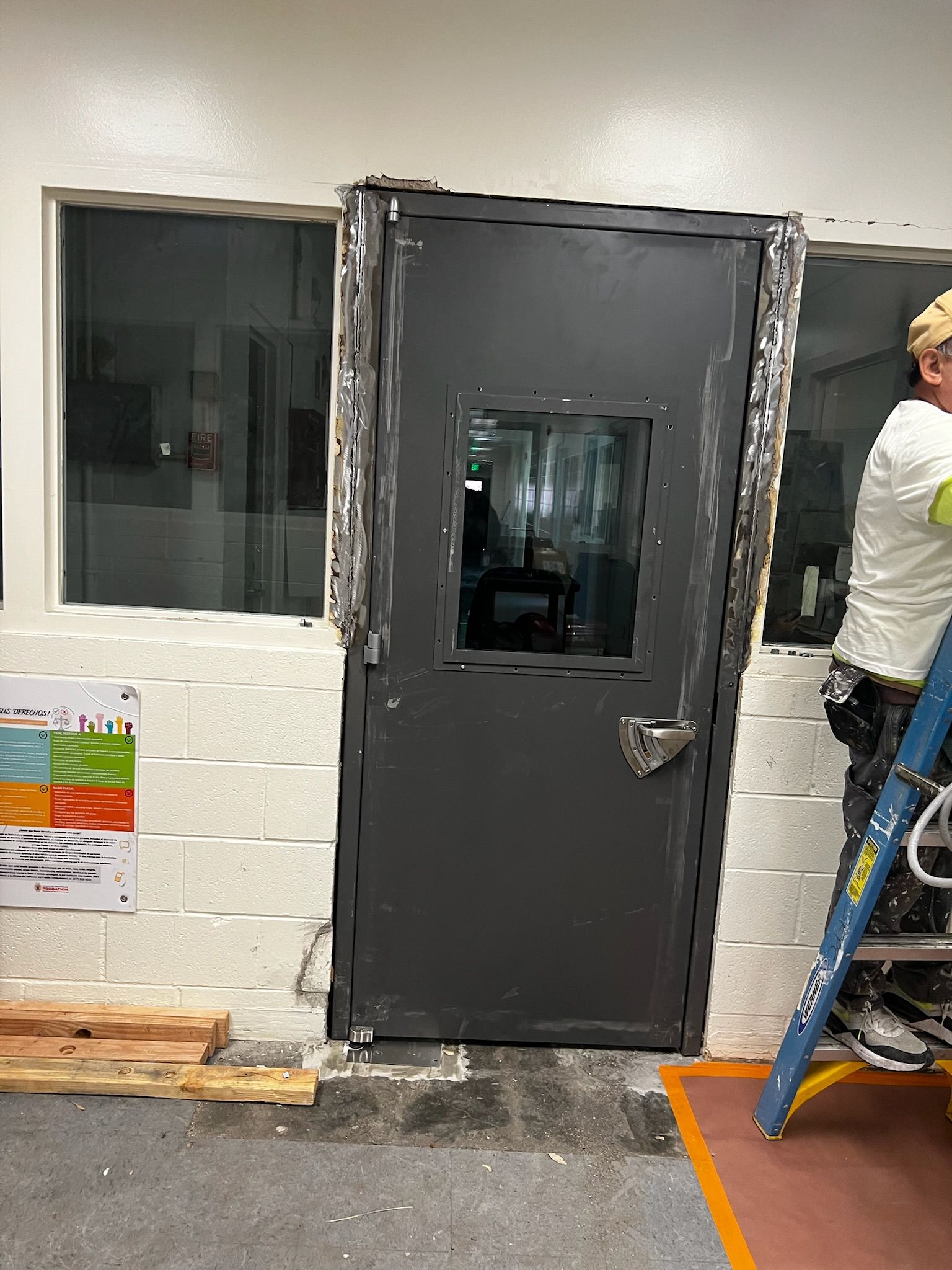 Dark gray door with window, set into brick and windowed wall. A person on a ladder is working on the side.