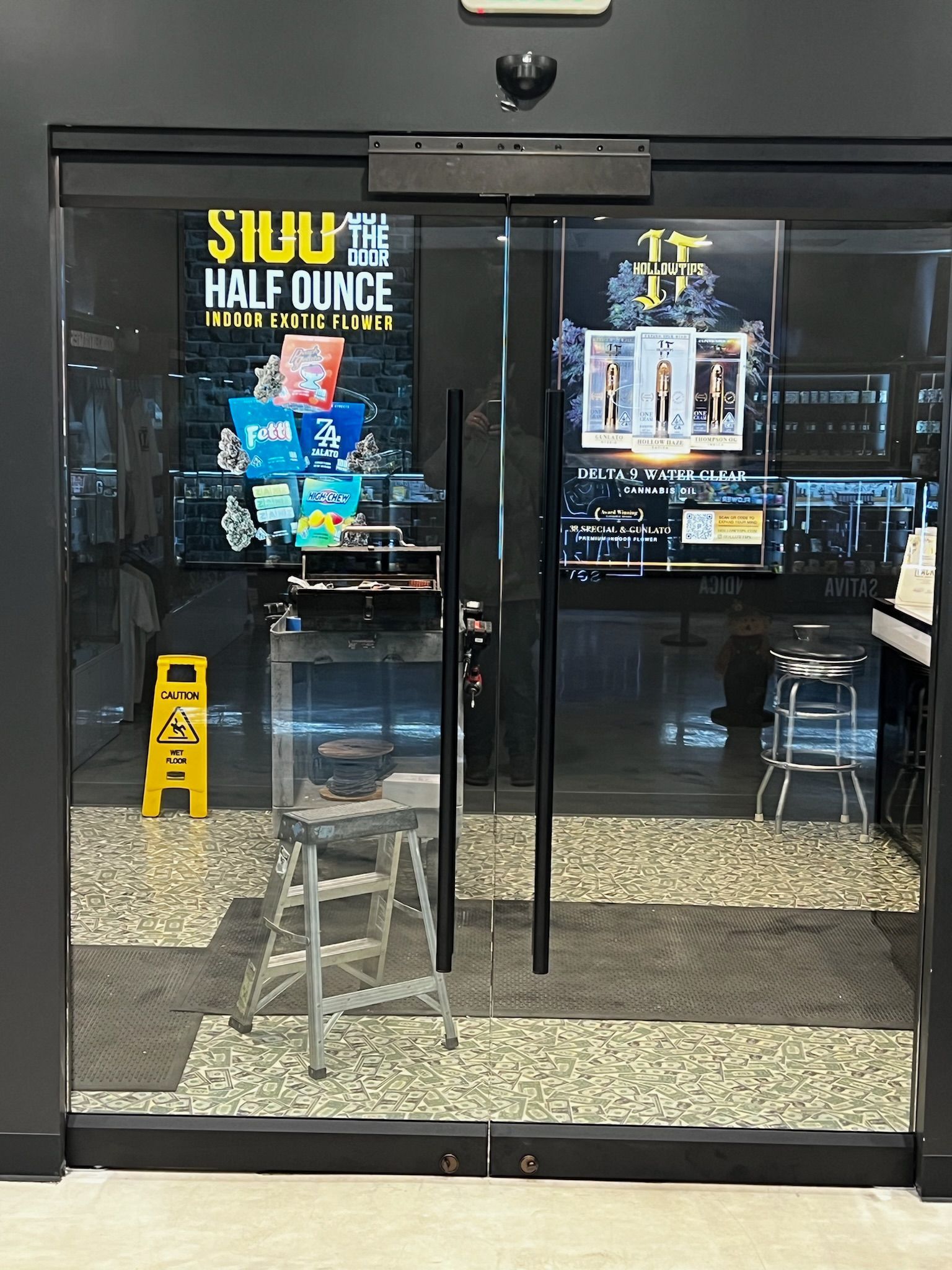 Glass doors to a store with advertising posters. A wet floor sign and a step stool are visible.