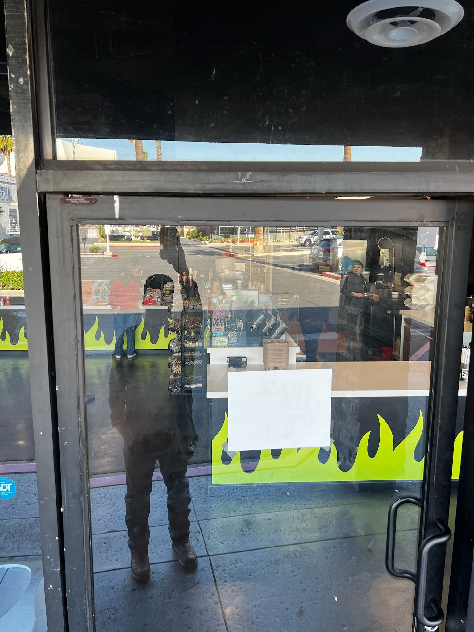 Person reflected in glass door, reaching up. Inside a store with a street visible through the glass.