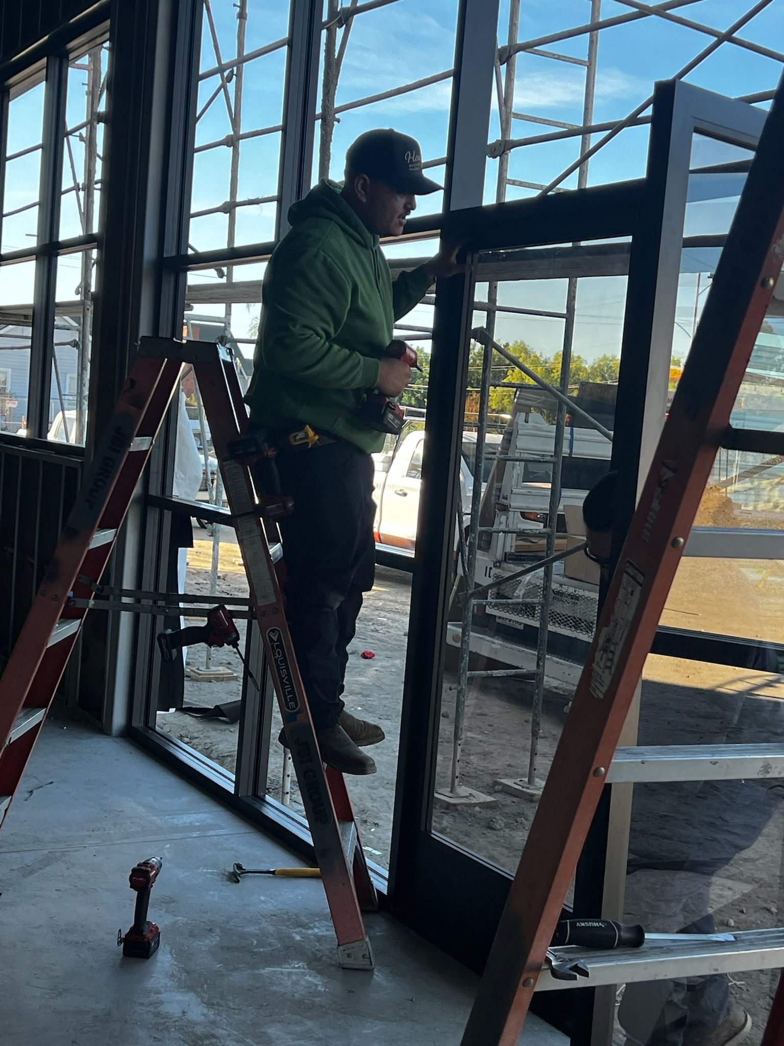 A worker in a green jacket on a ladder installs a window. The setting appears to be a construction site.