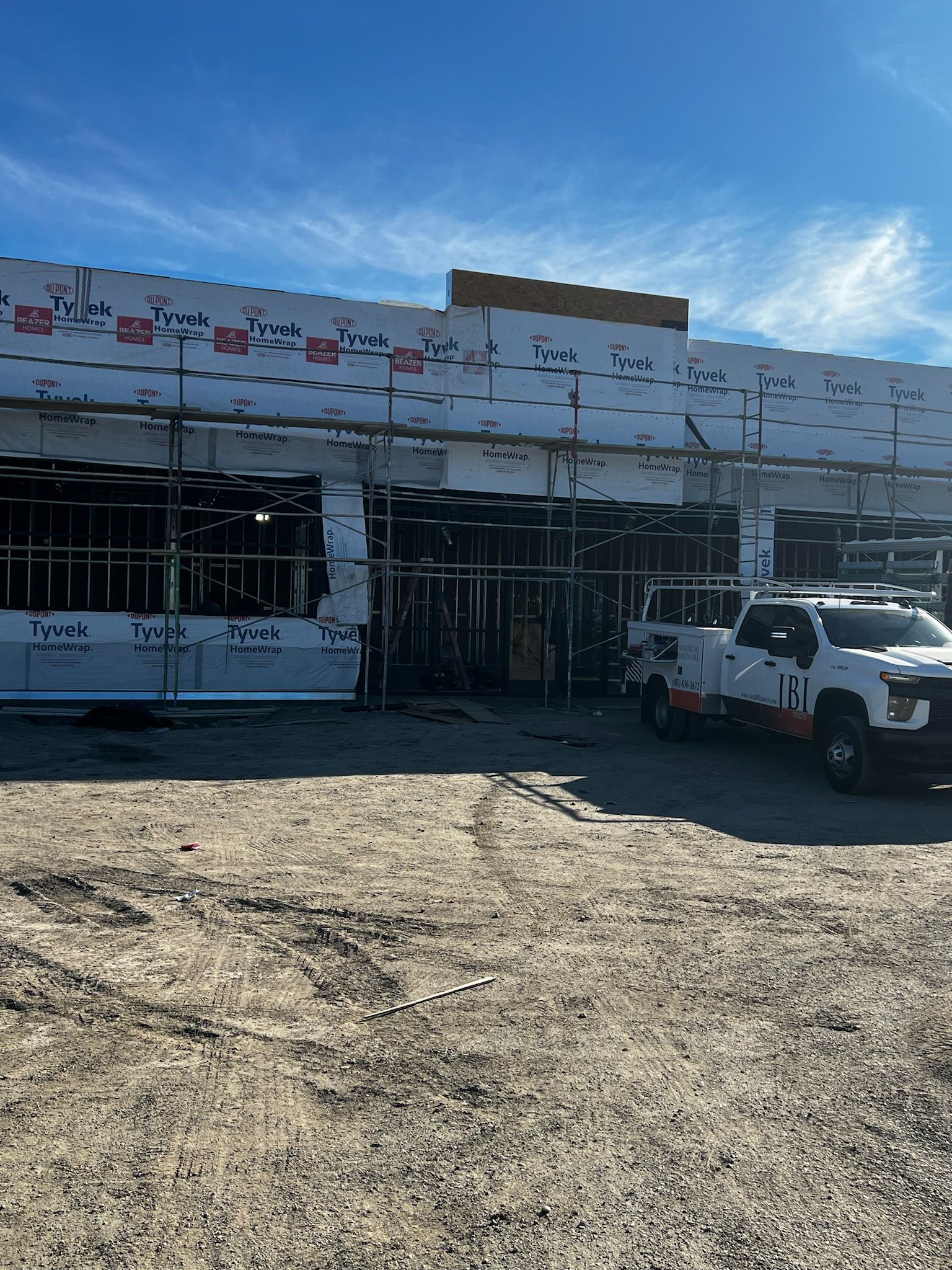 Building under construction with scaffolding and a work truck on a dirt lot, under a blue sky.