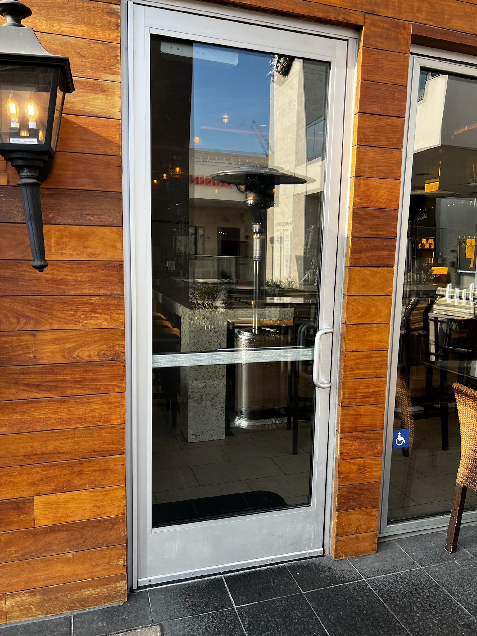 Glass door with silver frame, reflecting outdoor seating and heater. Adjacent to a wood-paneled wall, and a street lamp.