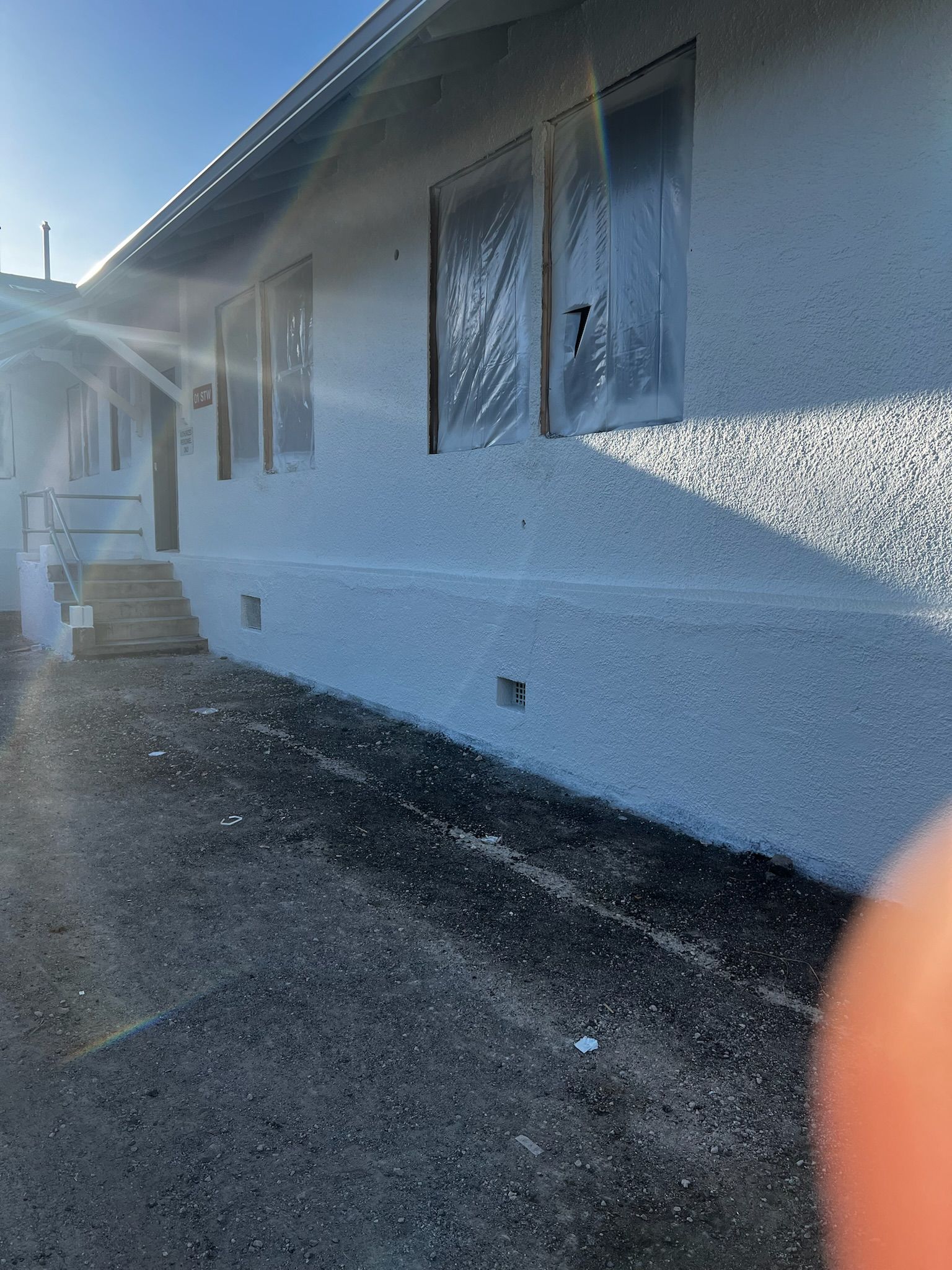 Exterior of a white stucco building with plastic-covered windows, sitting on a concrete surface.