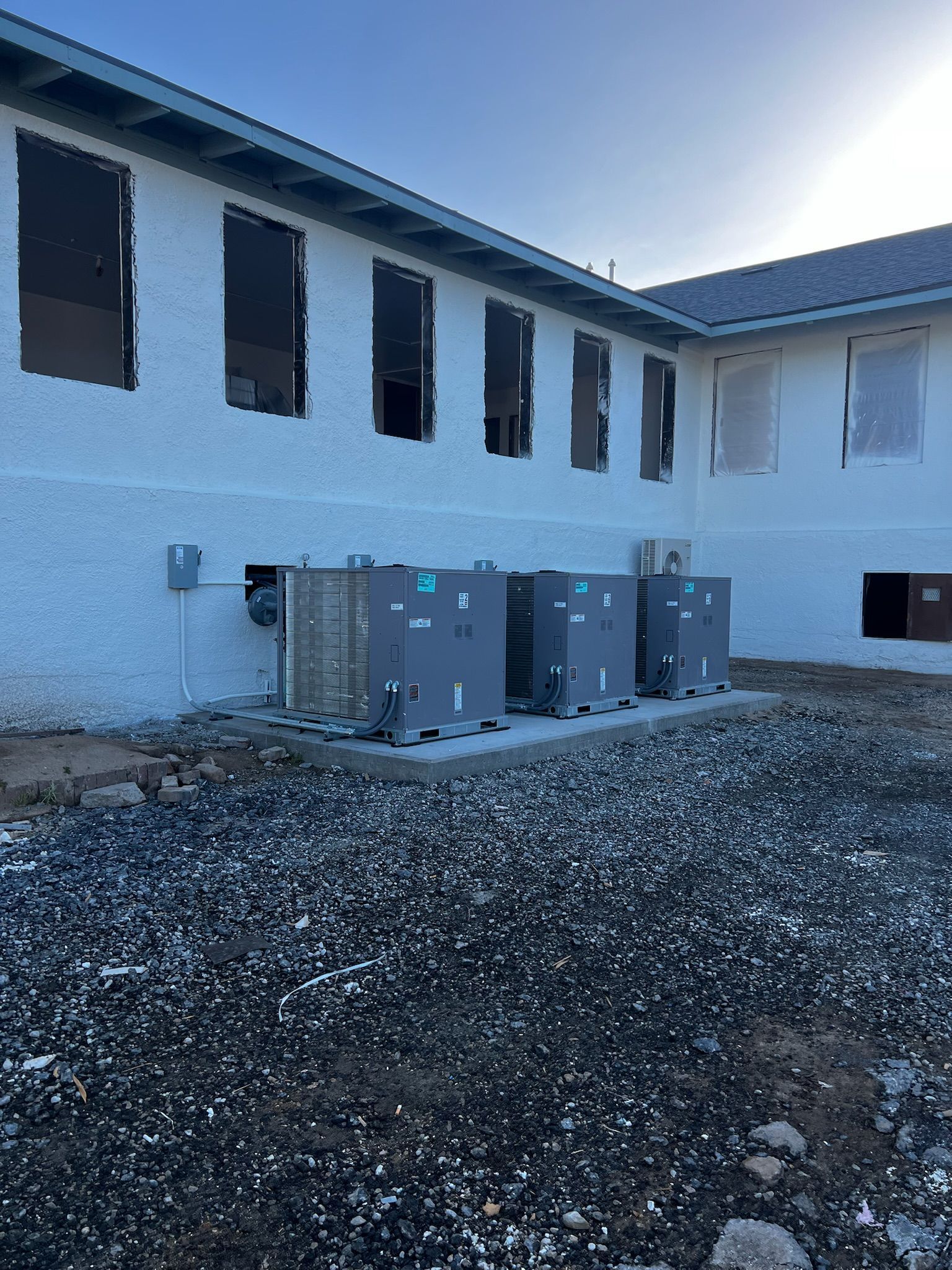Exterior of a white stucco building with multiple rectangular HVAC units on a gravel base. The windows are boarded up.