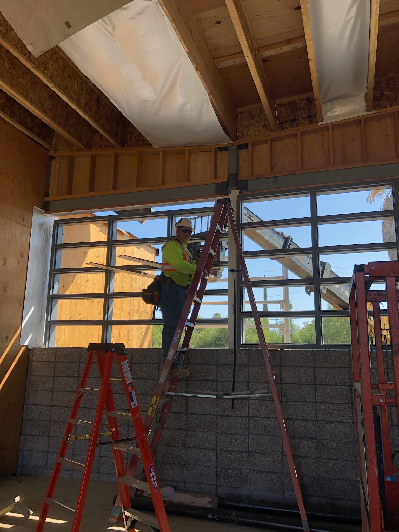 Construction worker on a ladder installing a glass window. Construction site with wooden beams and exterior visible.