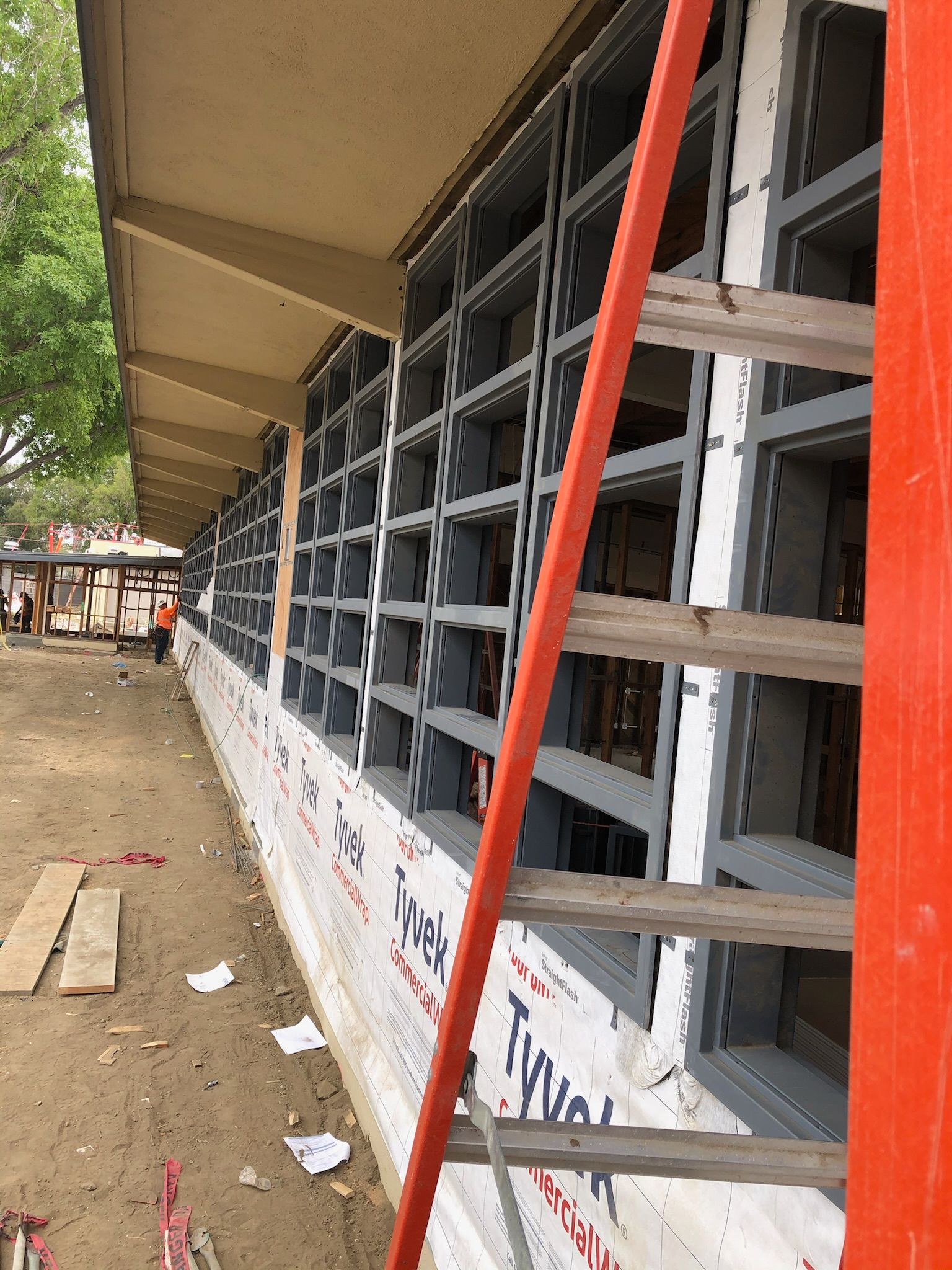 Building undergoing construction; window frames installed, ladder leaning against the wall, Tyvek wrap visible.