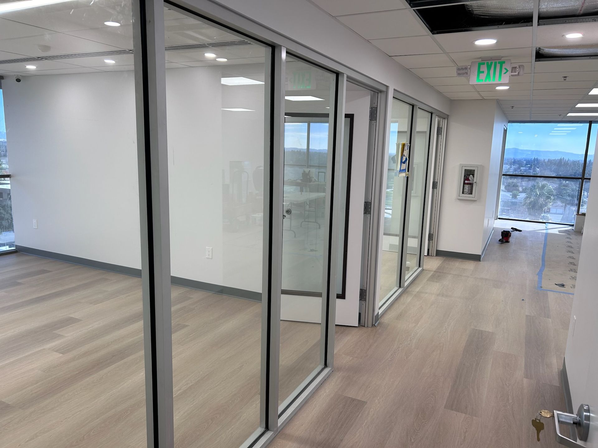 Empty office with glass-walled rooms, wood flooring, and an exit sign, with an outdoor view.