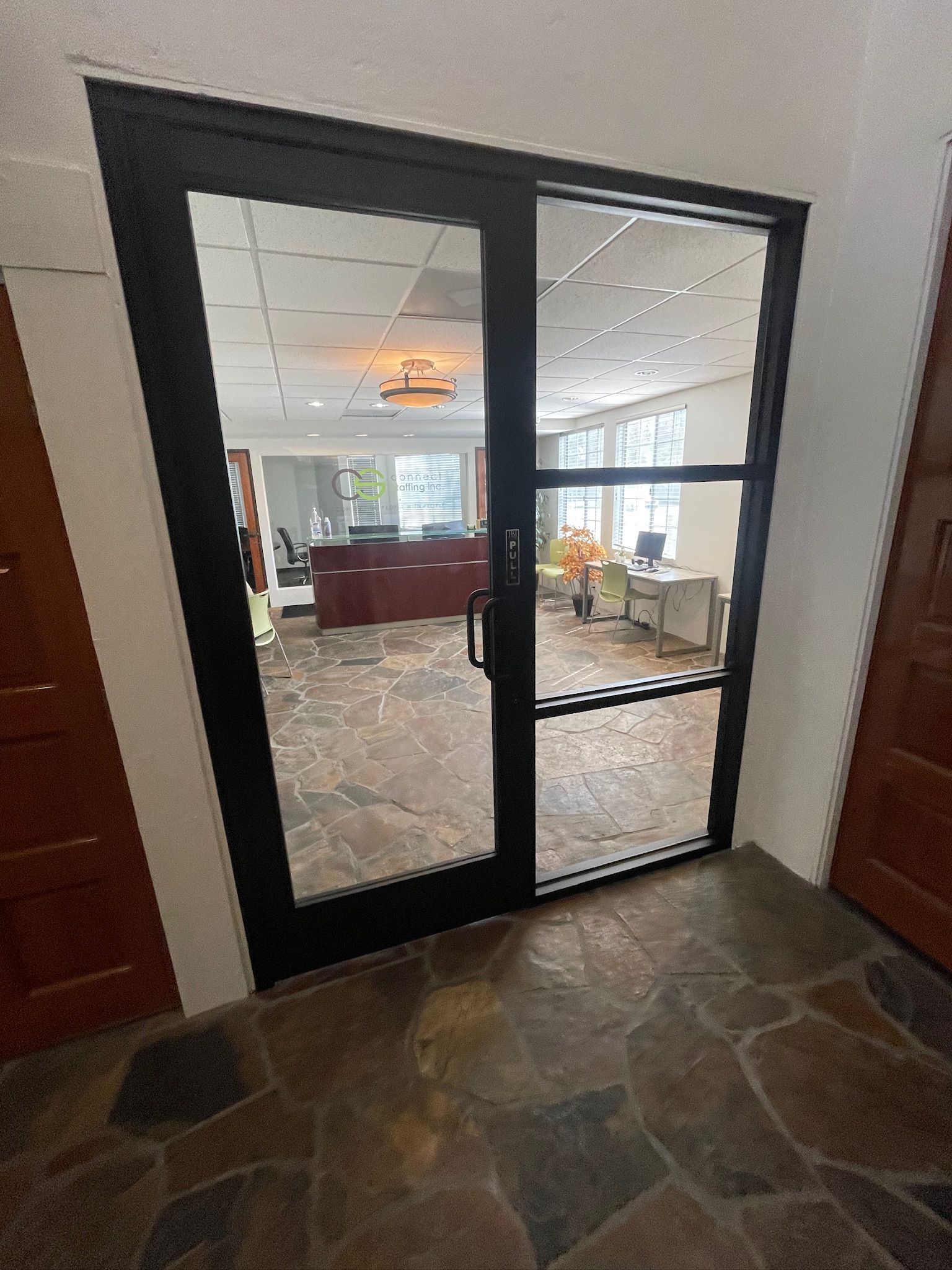 Black-framed glass doors open into an office space. Flooring is brown stone. A hallway is visible on either side.