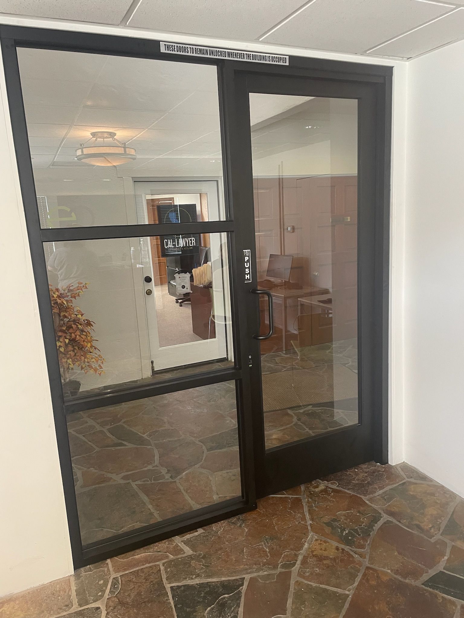 Glass doors with black frames, open, leading to an office space with stone flooring.