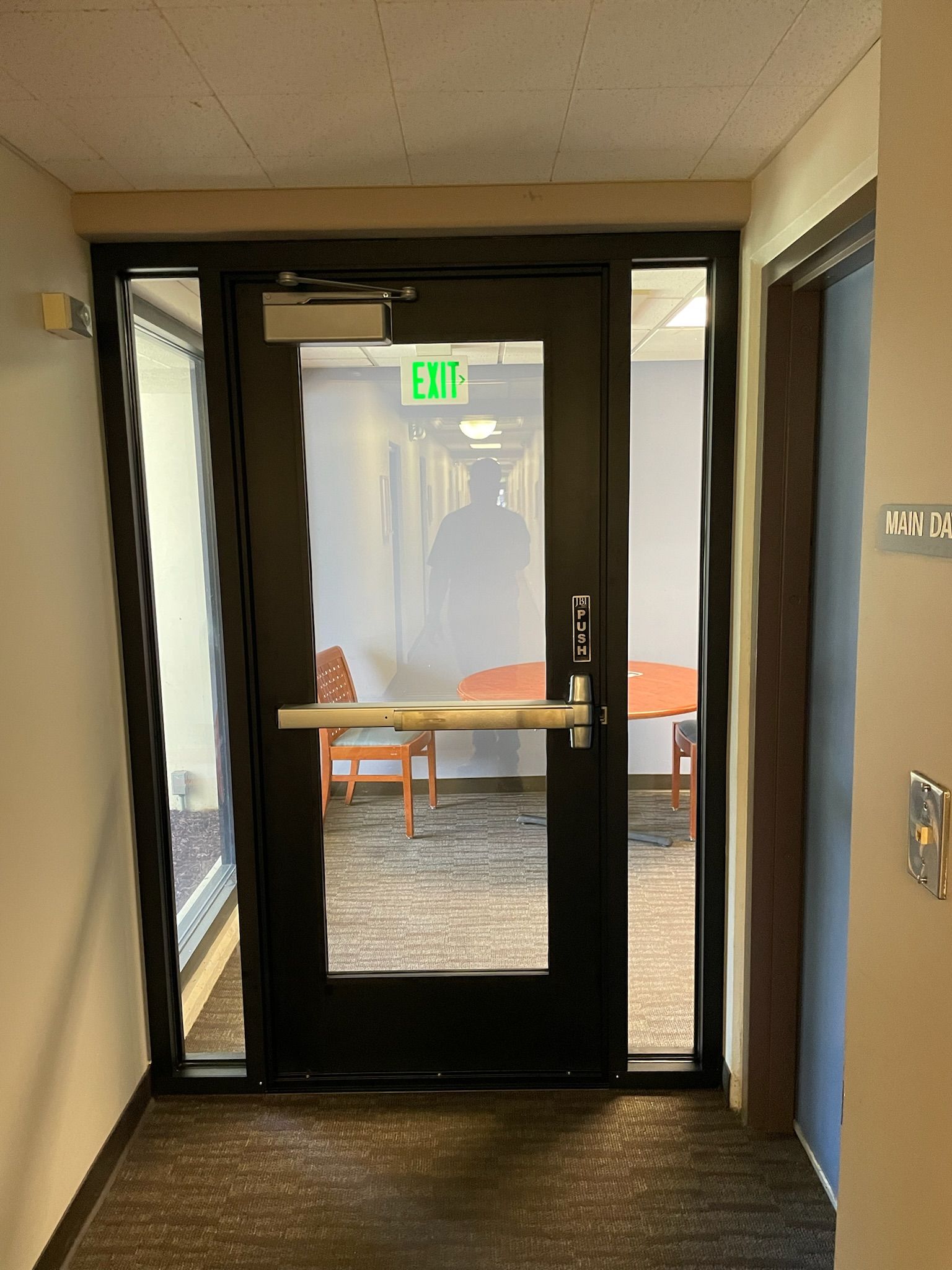 Black door with glass panel and exit sign; hallway with chairs visible through the door.