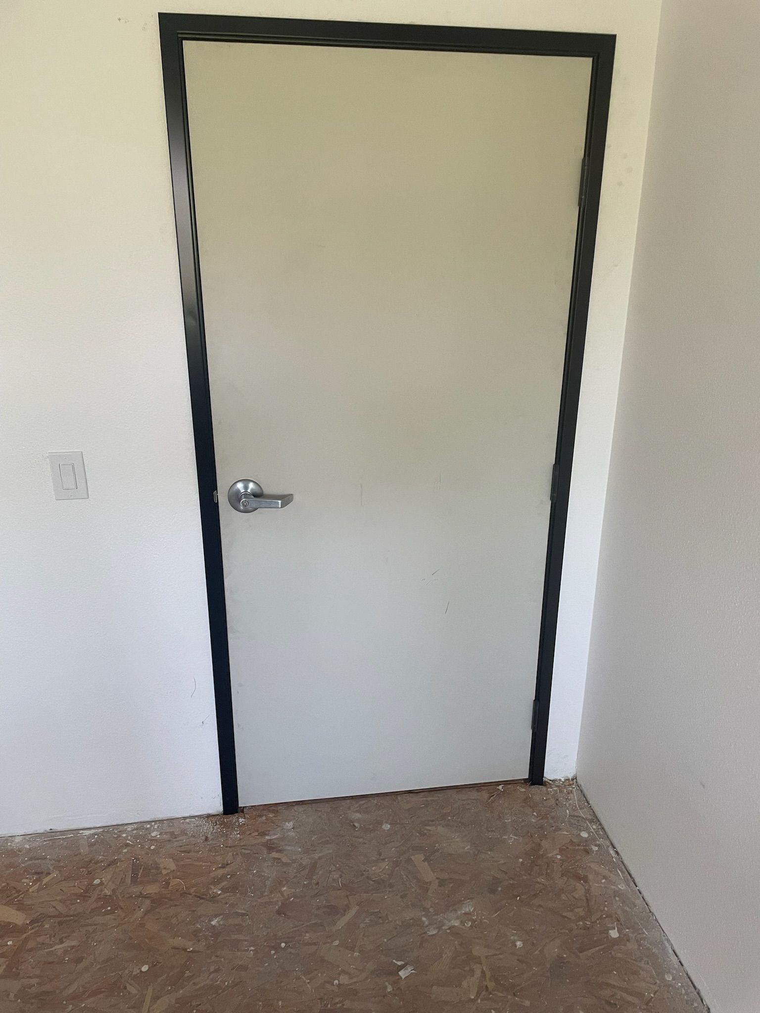 White door with black trim in a room with white walls and exposed flooring.