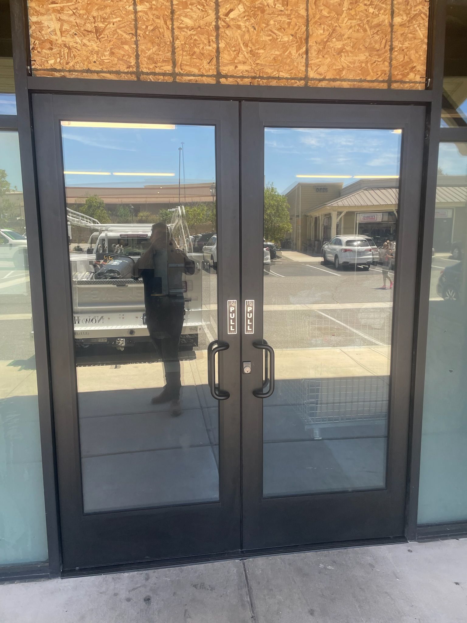 Double glass doors reflect a person and vehicles in an outdoor setting. Brown trim and a textured wood header.