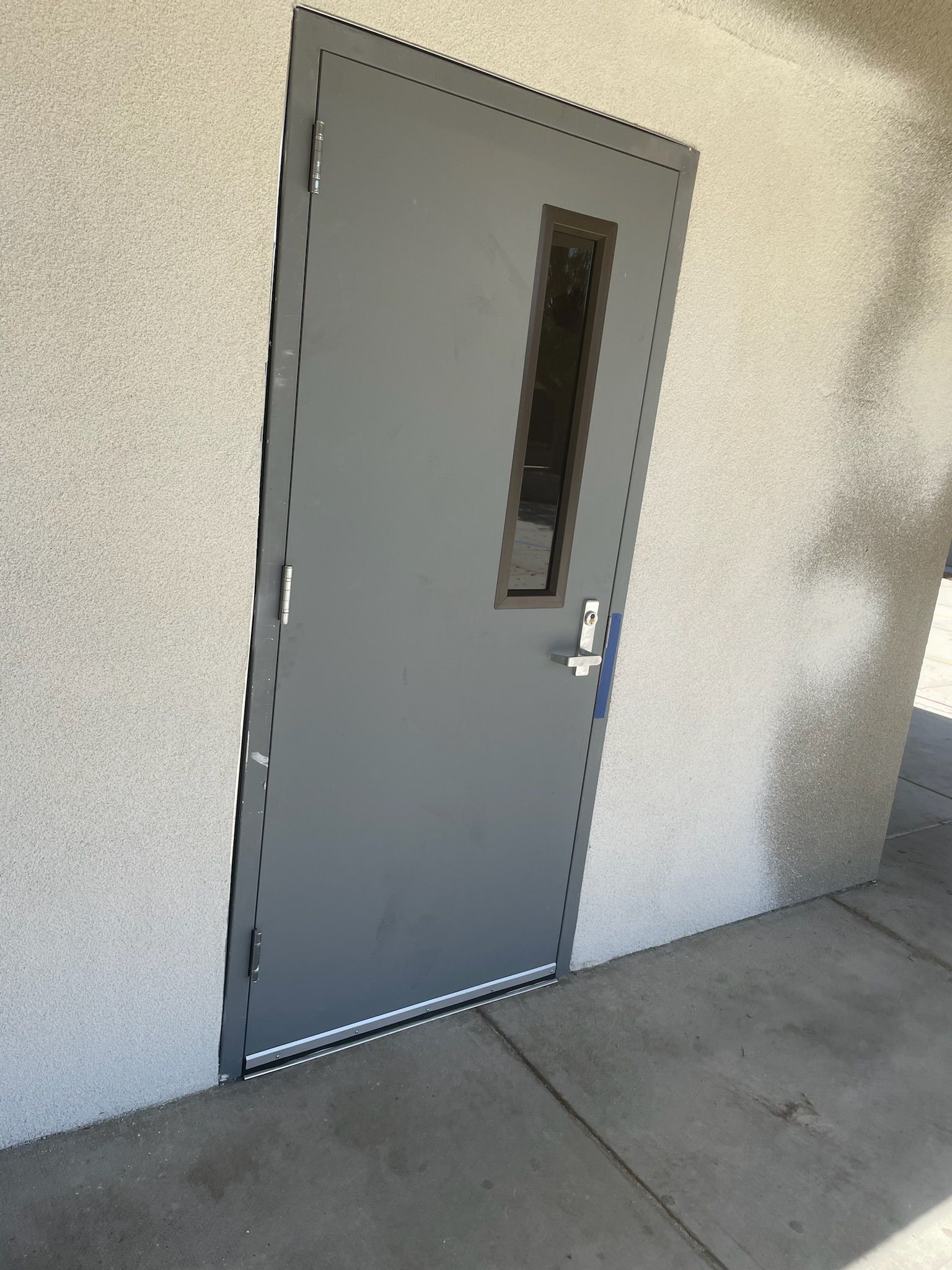 Gray metal door with a rectangular window and silver handle, set in a light stucco wall.