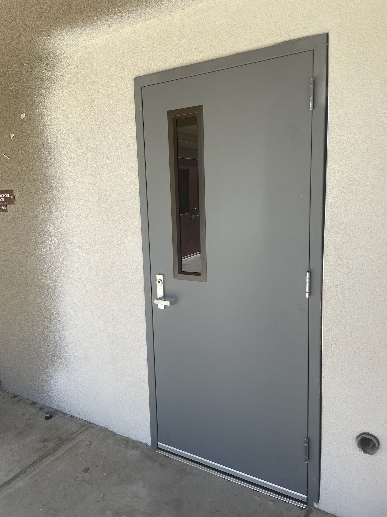 Gray metal door with small window set in a stucco wall.