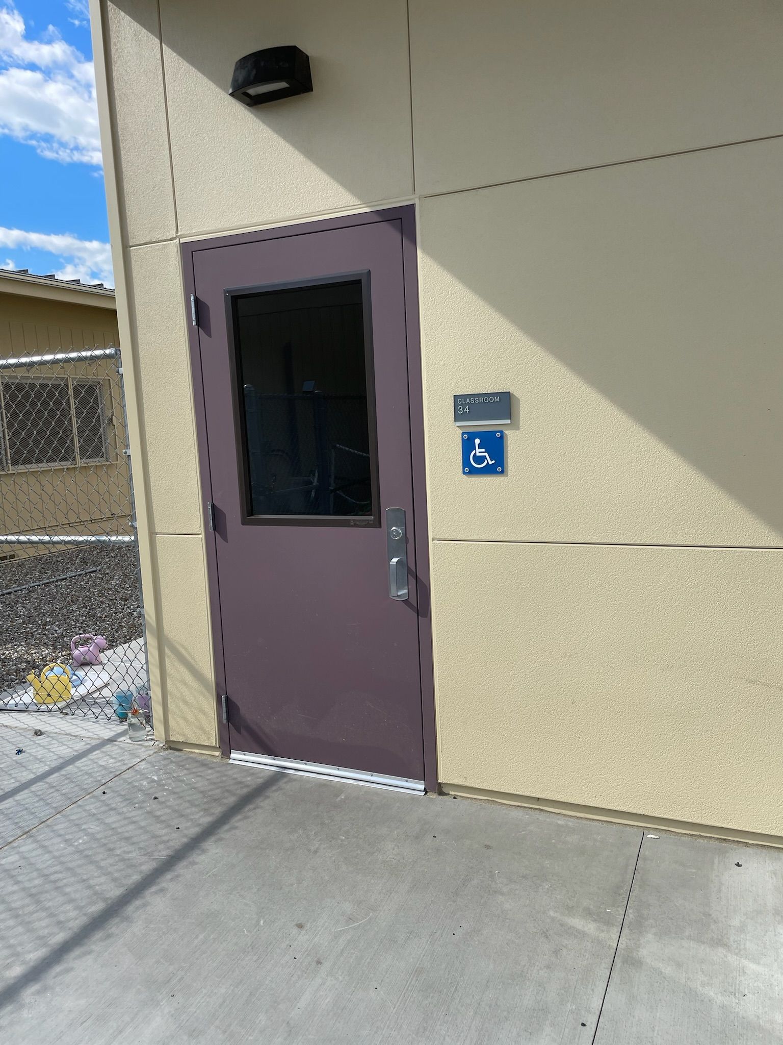 A tan building with a brown door, window, and accessible sign. Shadowed sidewalk, chain-link fence.