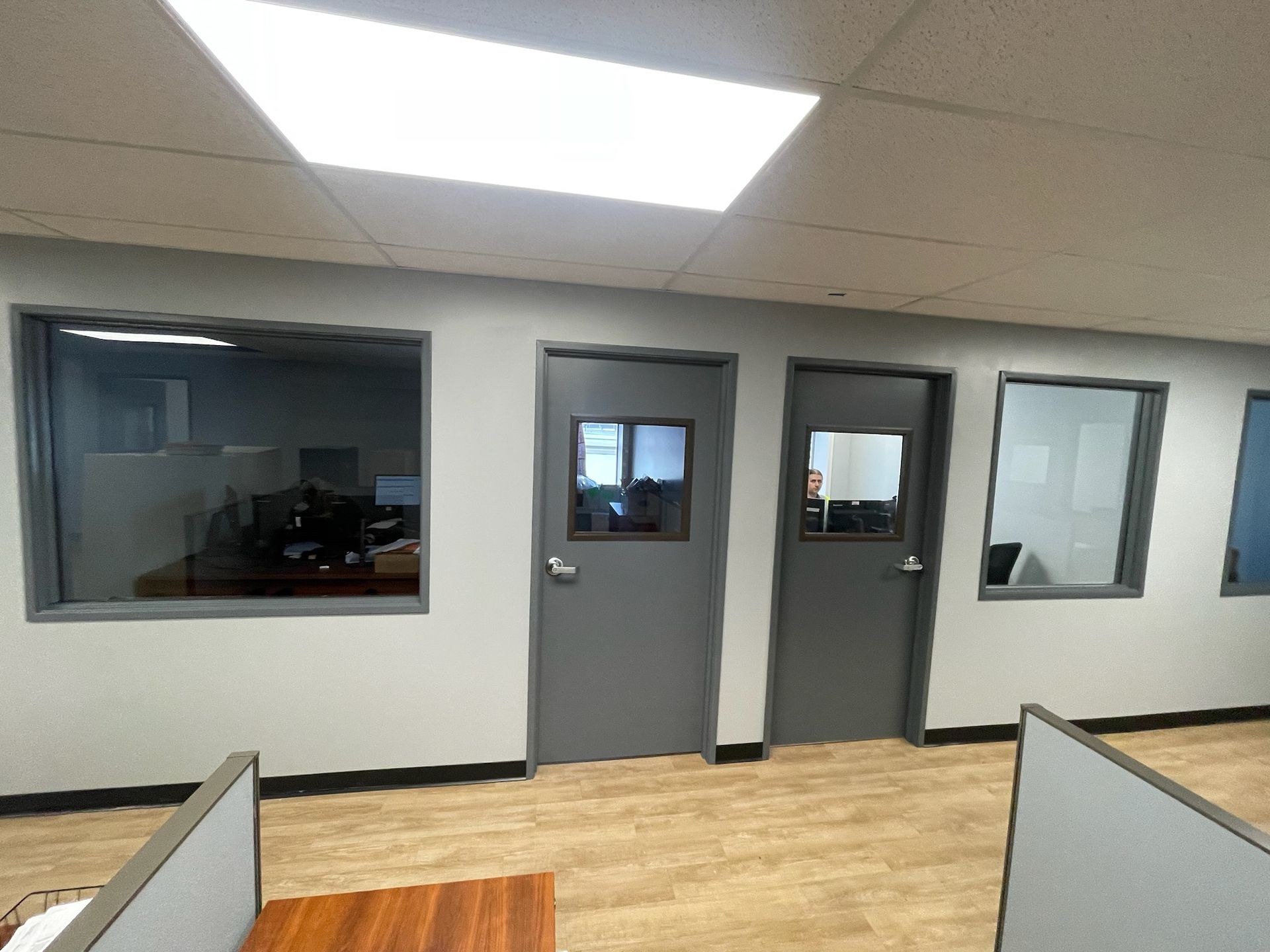 Office interior with grey doors, windows, and cubicle dividers. Fluorescent lighting.