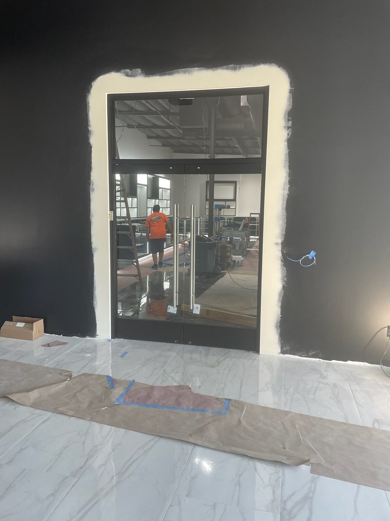 Doorway with clear glass doors; person in orange shirt reflected in the glass, black wall, white trim, tile floor.