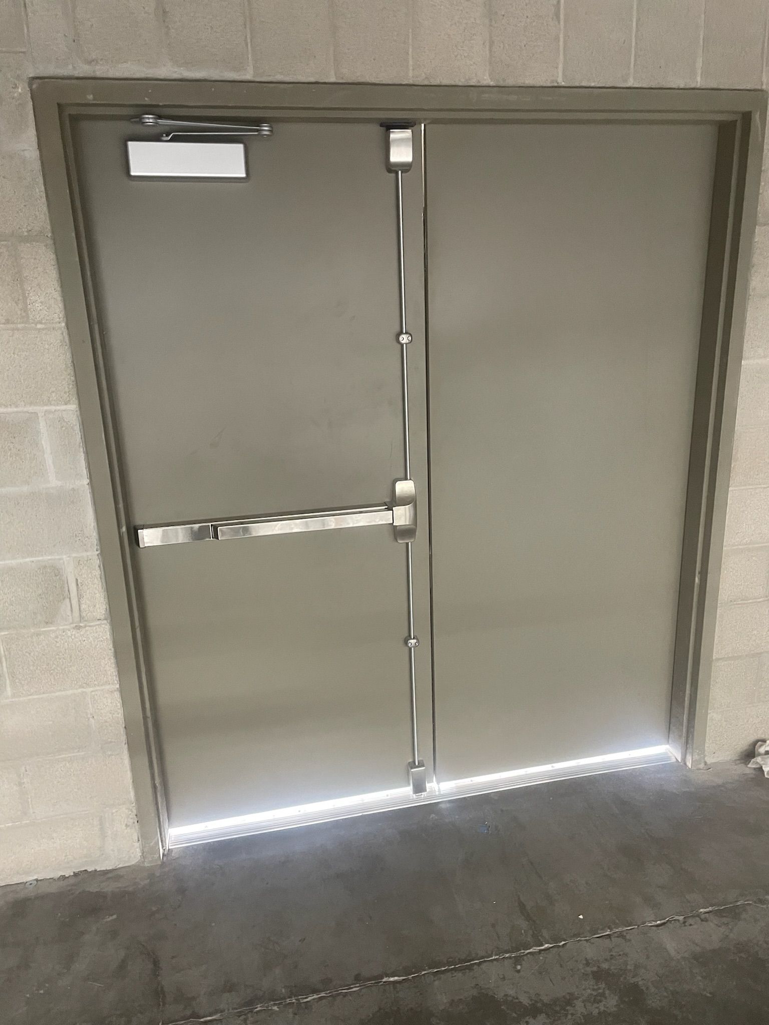 Closed gray double door with horizontal bar, in a cinder block wall.