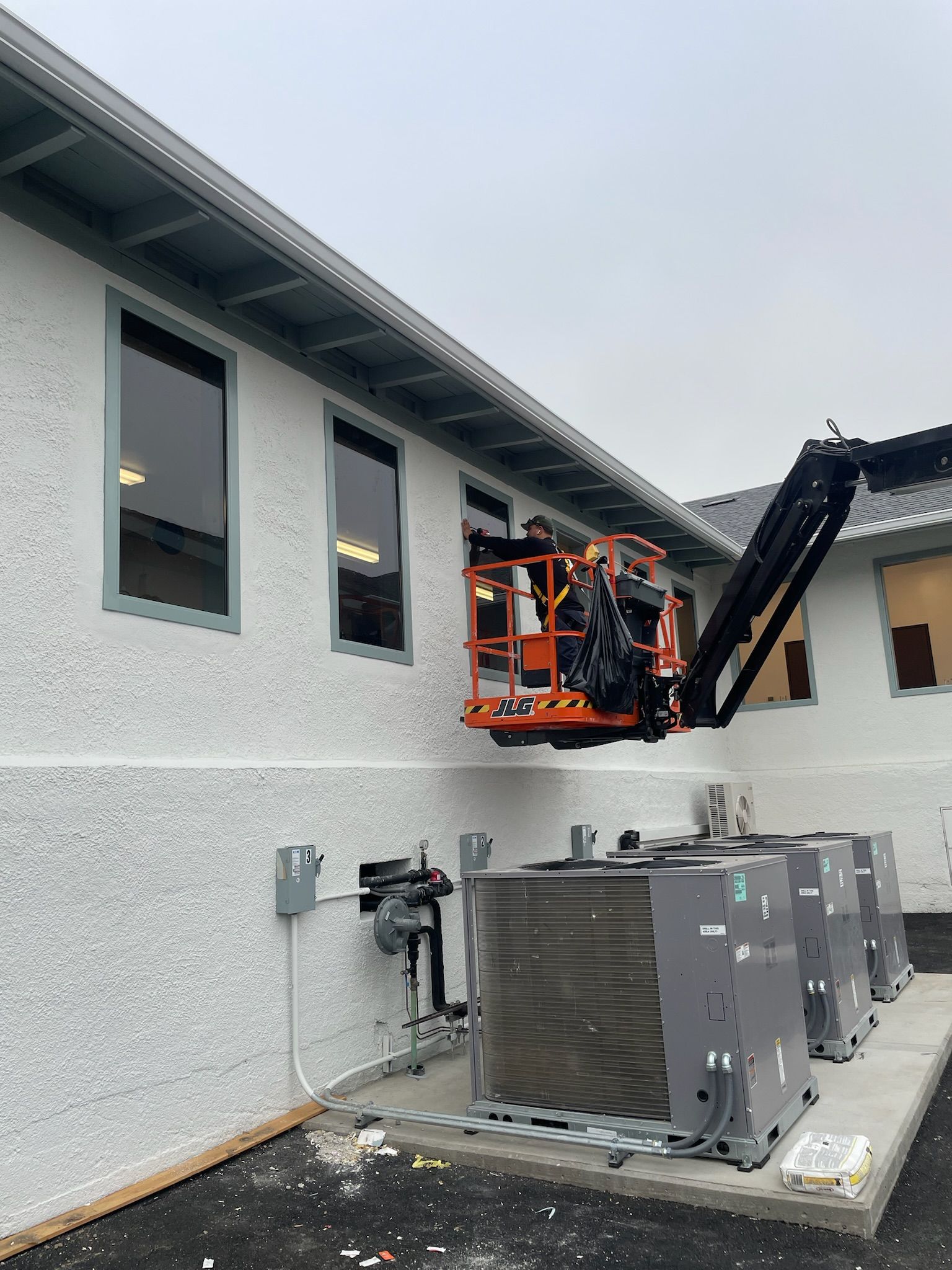 Workers in an aerial lift near a building's windows, possibly for maintenance. Gray sky.