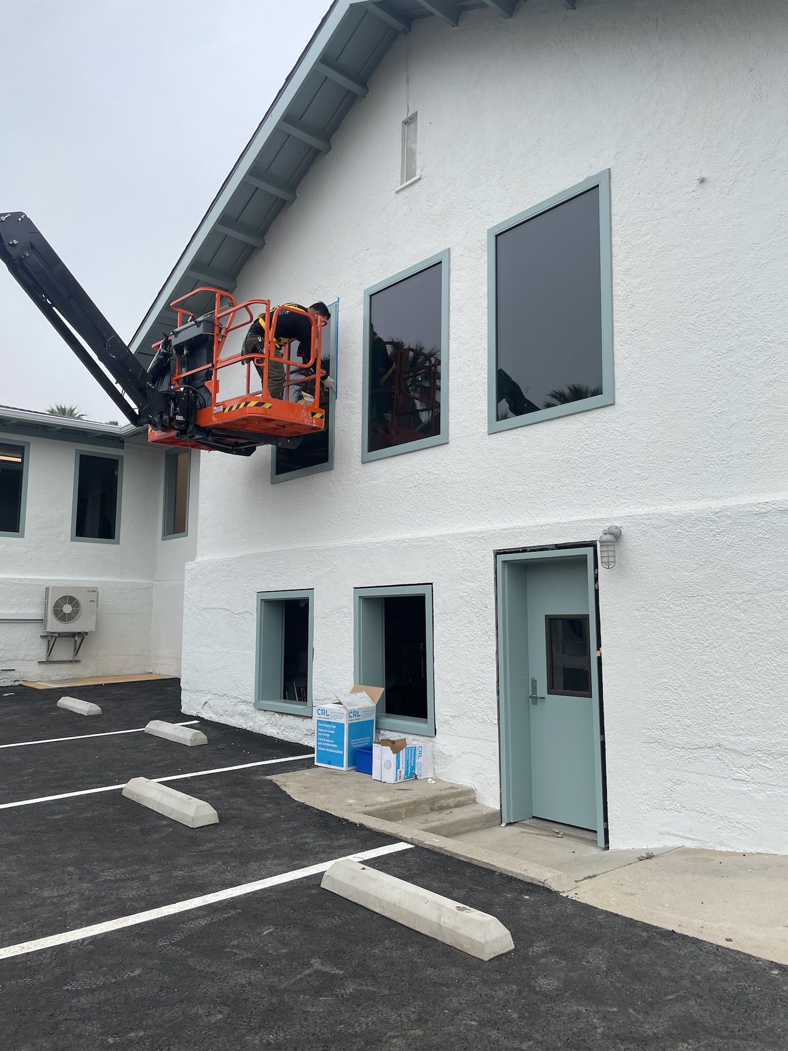 A person in an orange lift works on a white building, replacing windows.