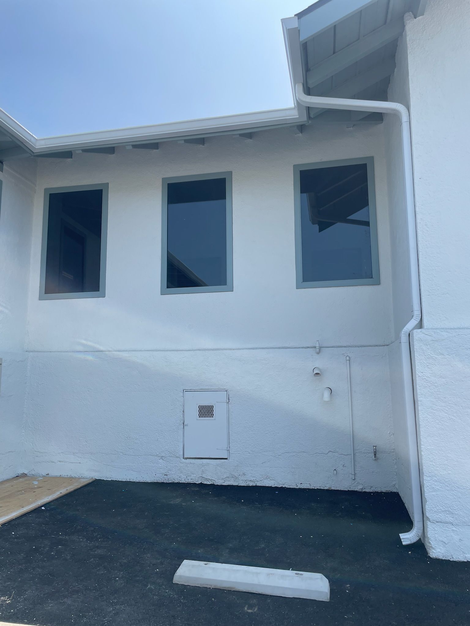 Exterior wall of a building with three windows and a small door, painted white with blue trim, black asphalt below.