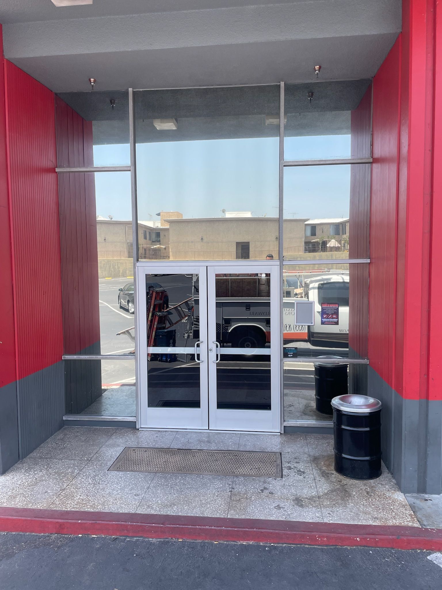 Glass double doors lead into a building with red and gray exterior, with a trash can on either side.