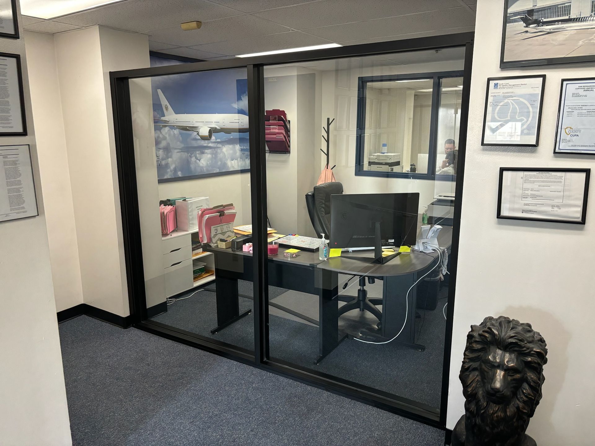 Office with glass walls, desk, computer, and decor; a lion statue sits on the right.