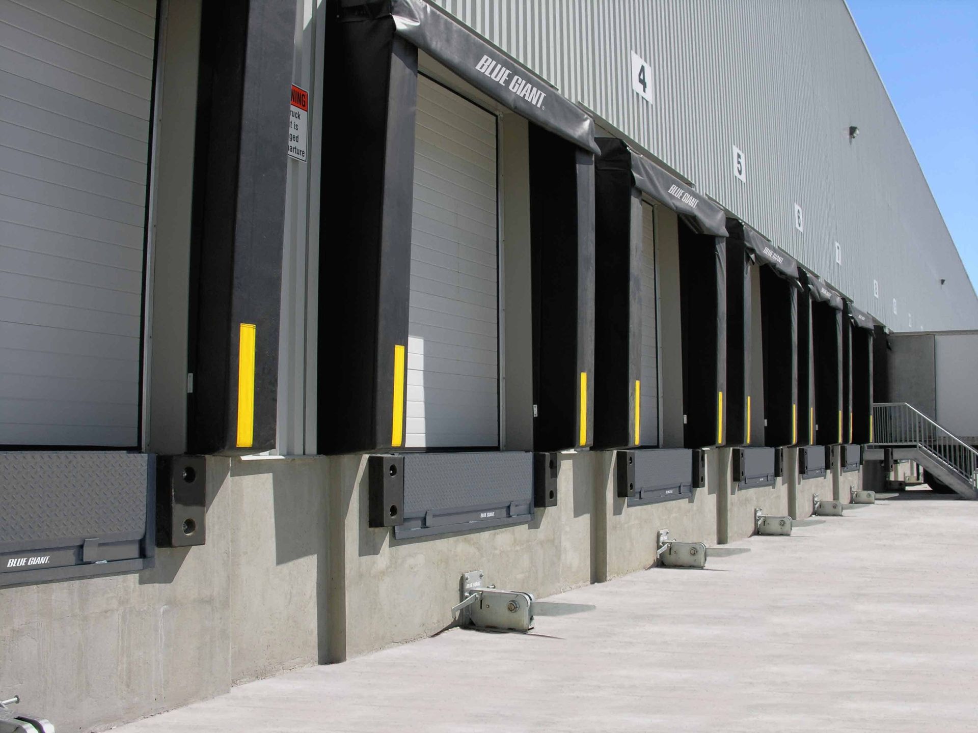 Loading docks on a warehouse exterior. Gray walls, black seals, white doors, concrete pad, blue sky.
