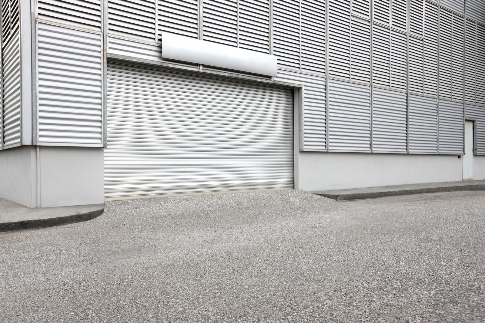 Exterior view of a building with a gray metallic roll-up door and a gravel driveway.