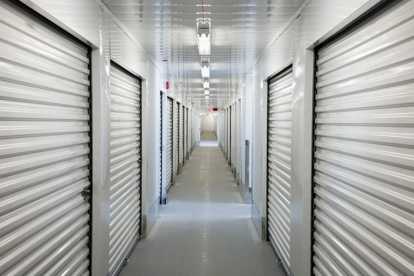 Hallway of storage units with white, roll-up doors and fluorescent lights.