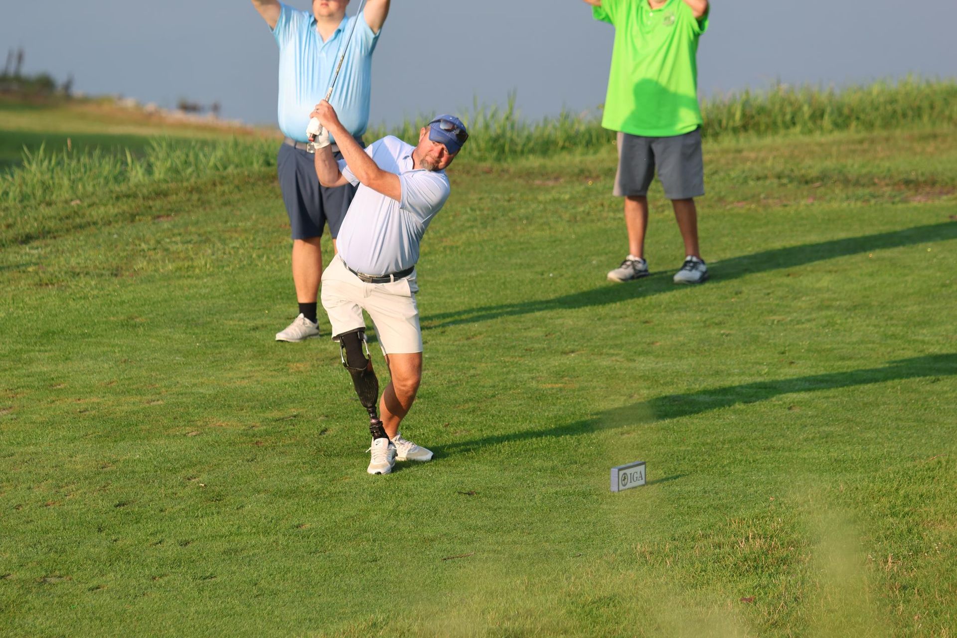 Golfer with prosthetic leg swings club on green; two men cheer behind him.